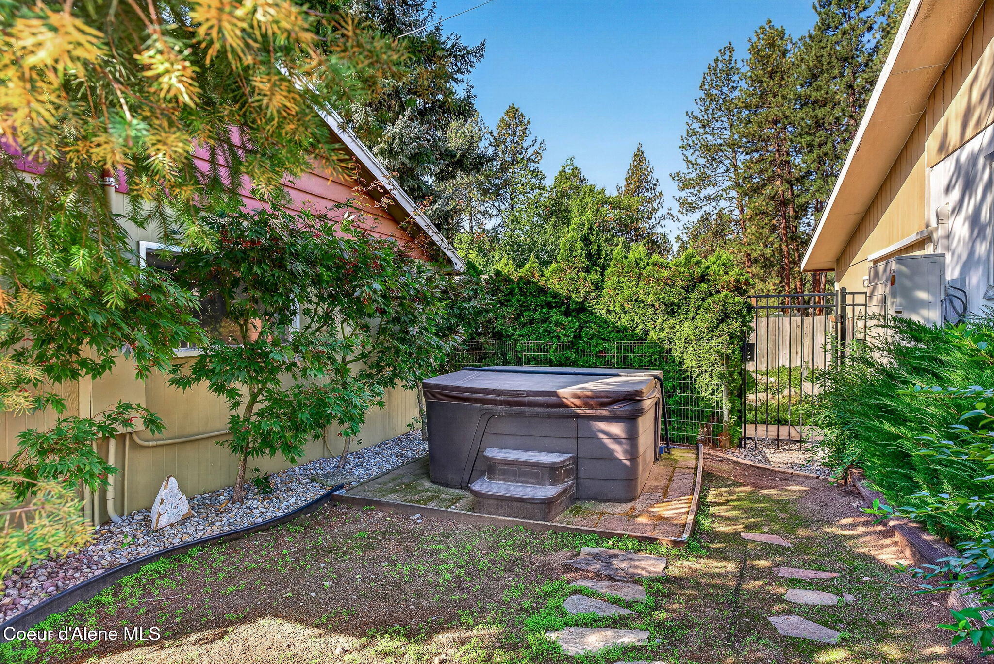Outdoor hot tub with steps in a landscaped, fenced yard between buildings.