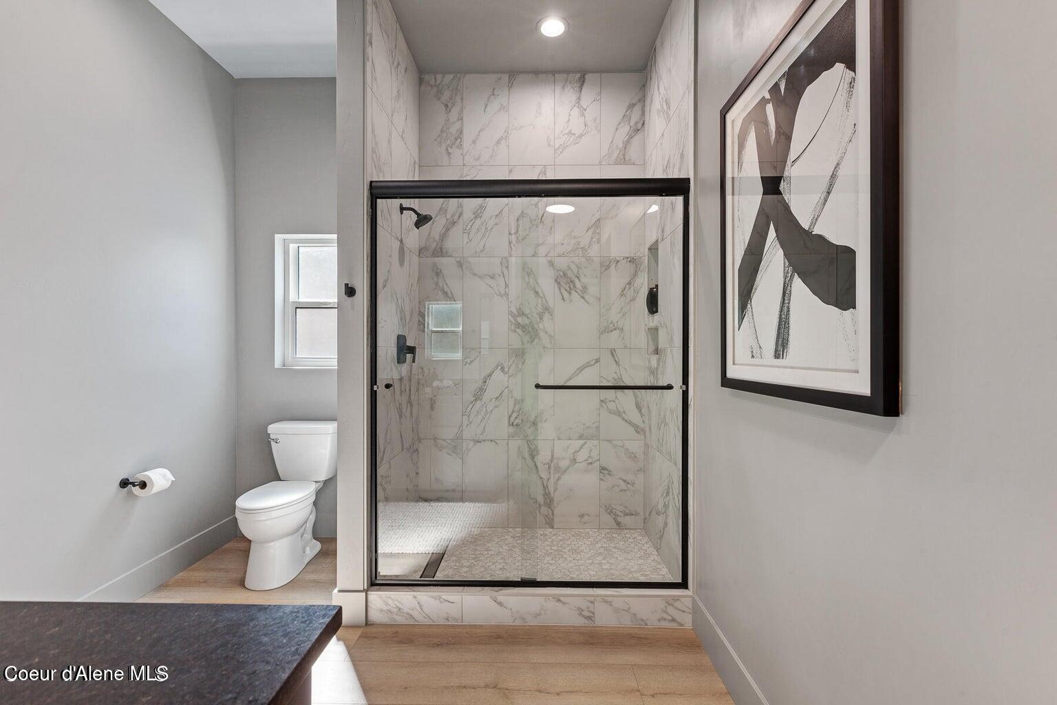 Modern bathroom with a marble-look glass shower, black hardware, and a toilet beside a small window.