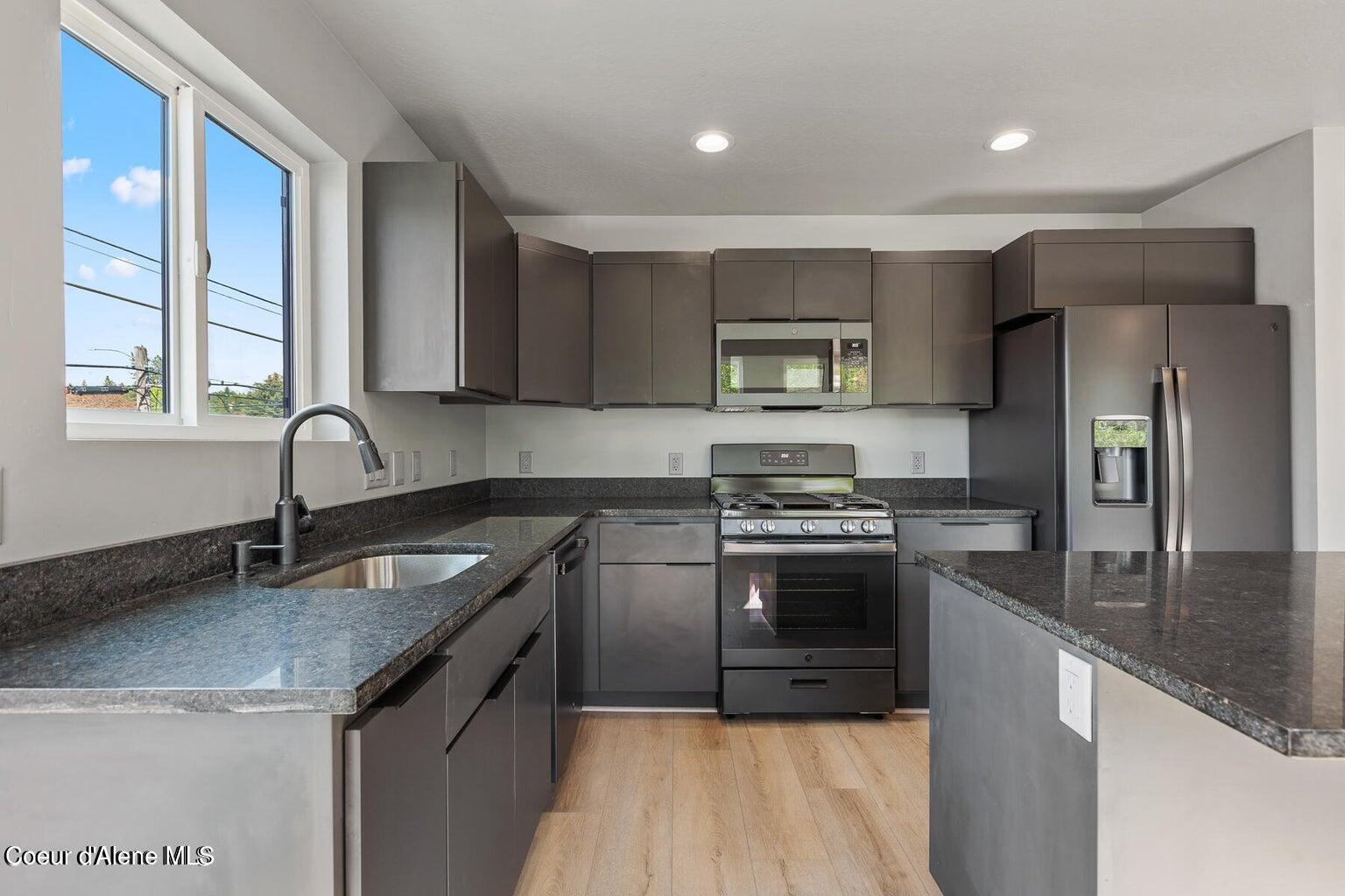 Modern kitchen with dark cabinets, stone counters, stainless steel appliances, and a large window.