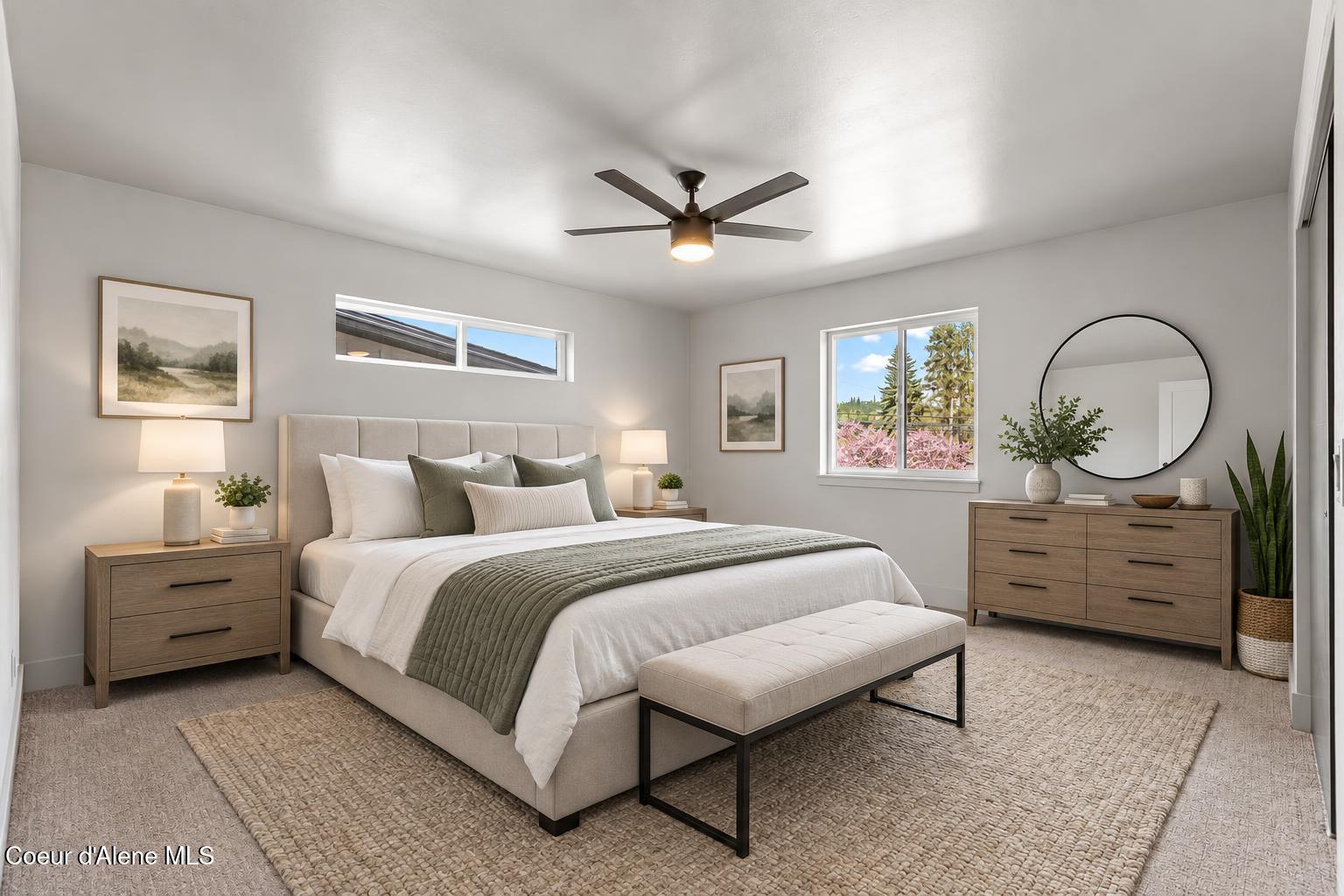 Staged bedroom with a large upholstered bed, sage-green throw, wood furniture, and windows overlooking trees.