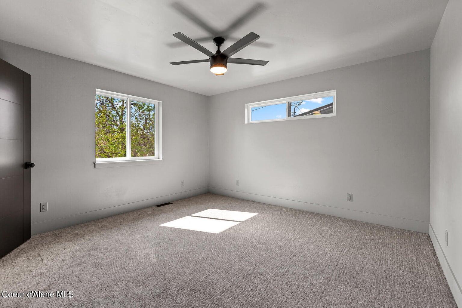 Empty bedroom with gray carpet, two windows, and a ceiling fan.