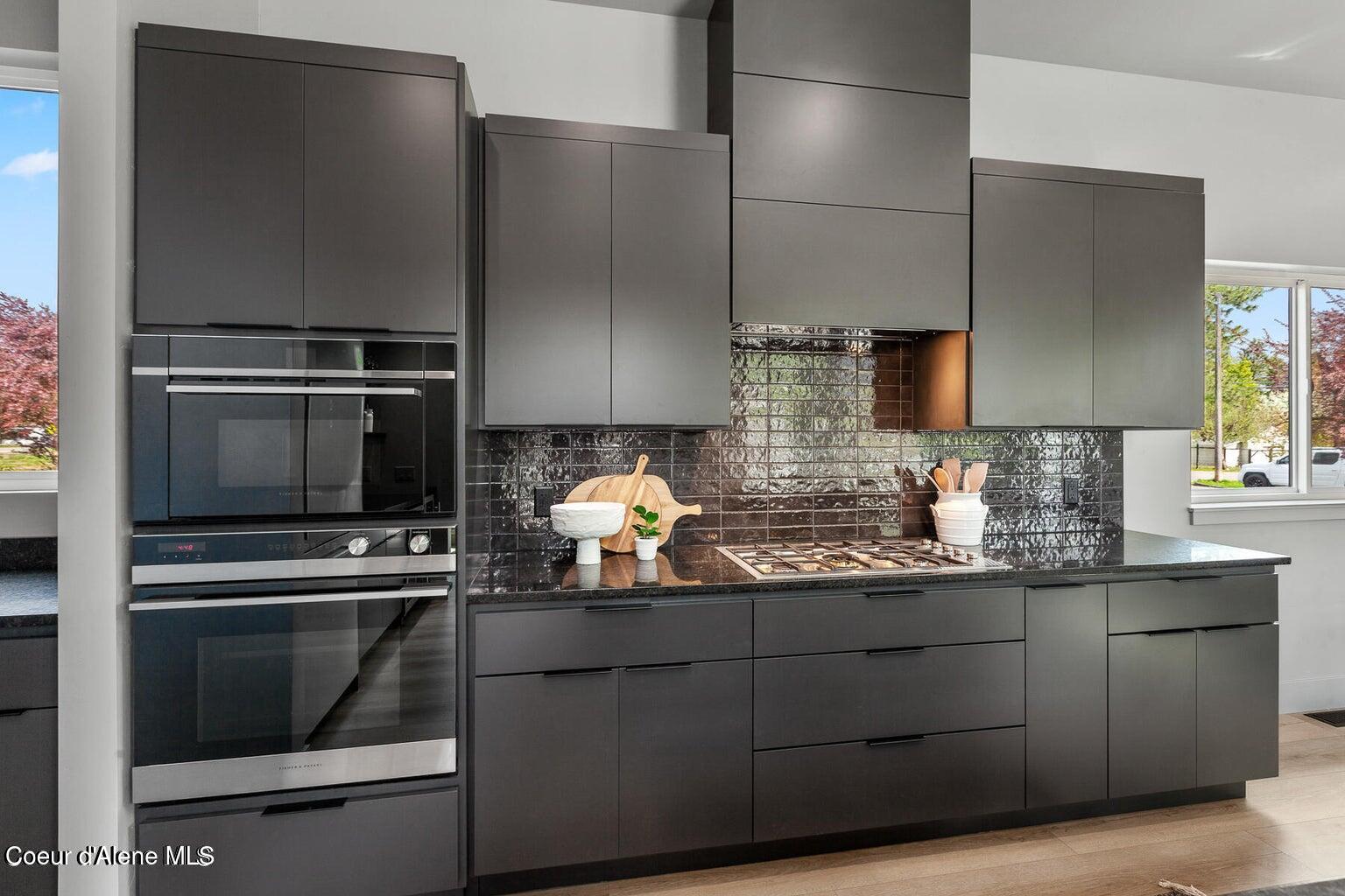 Modern kitchen with dark cabinetry, glossy tile backsplash, gas cooktop, and stacked built-in ovens.