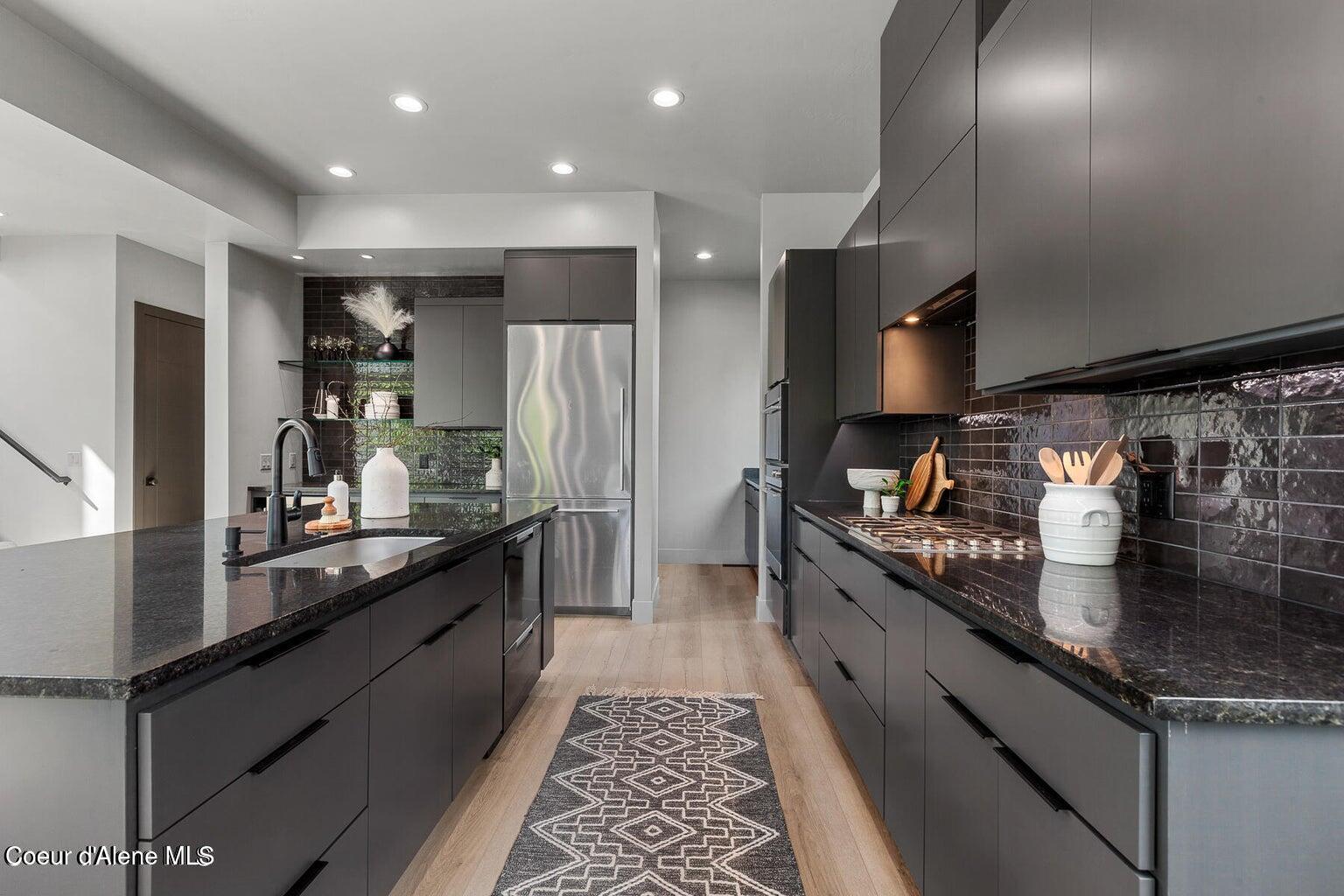 Modern kitchen with dark cabinetry, black stone counters, stainless steel refrigerator, and a patterned runner rug.