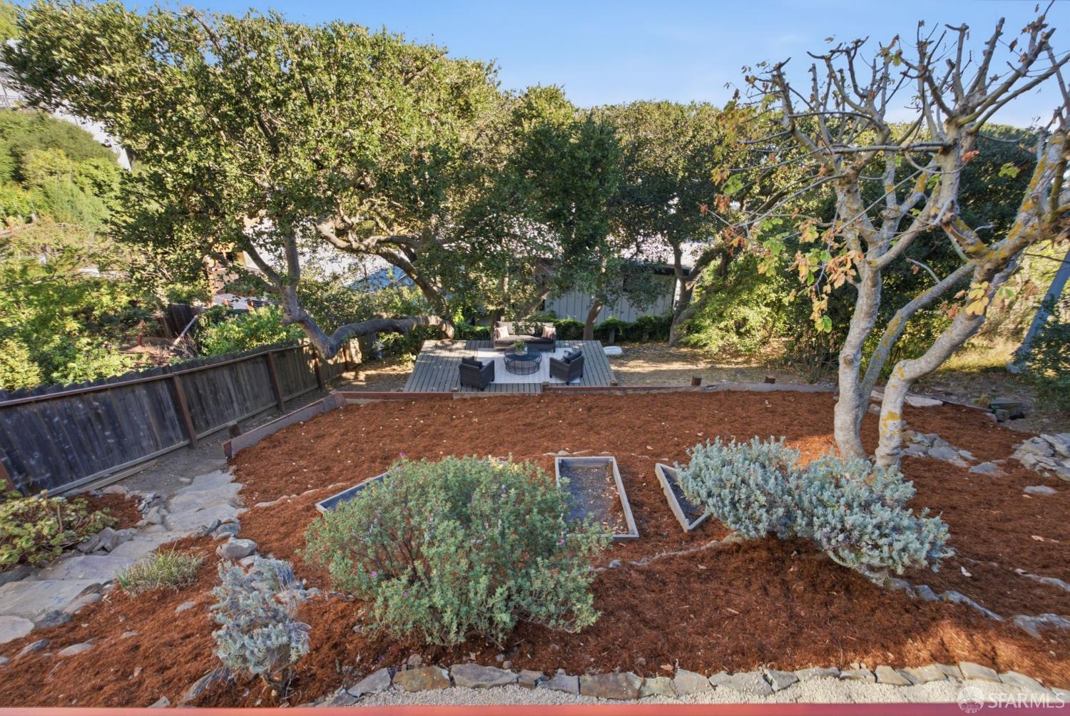 Backyard garden with a raised deck, outdoor seating, mature trees, and mulched planting beds.