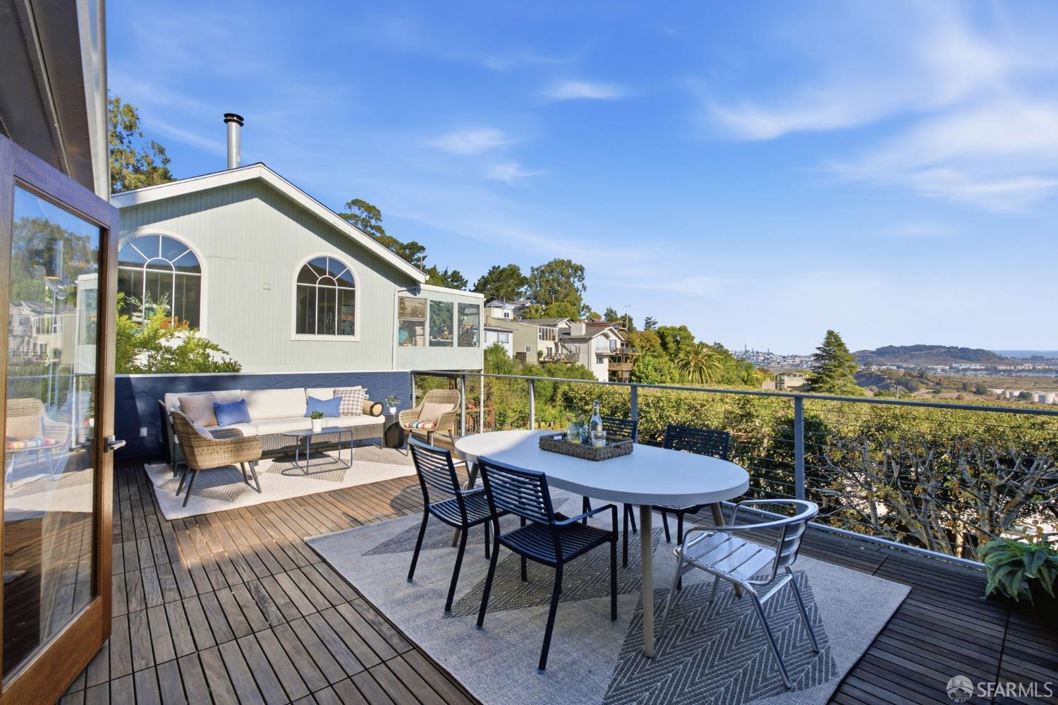 Furnished wooden deck with dining and lounge areas overlooking greenery and a distant bay.