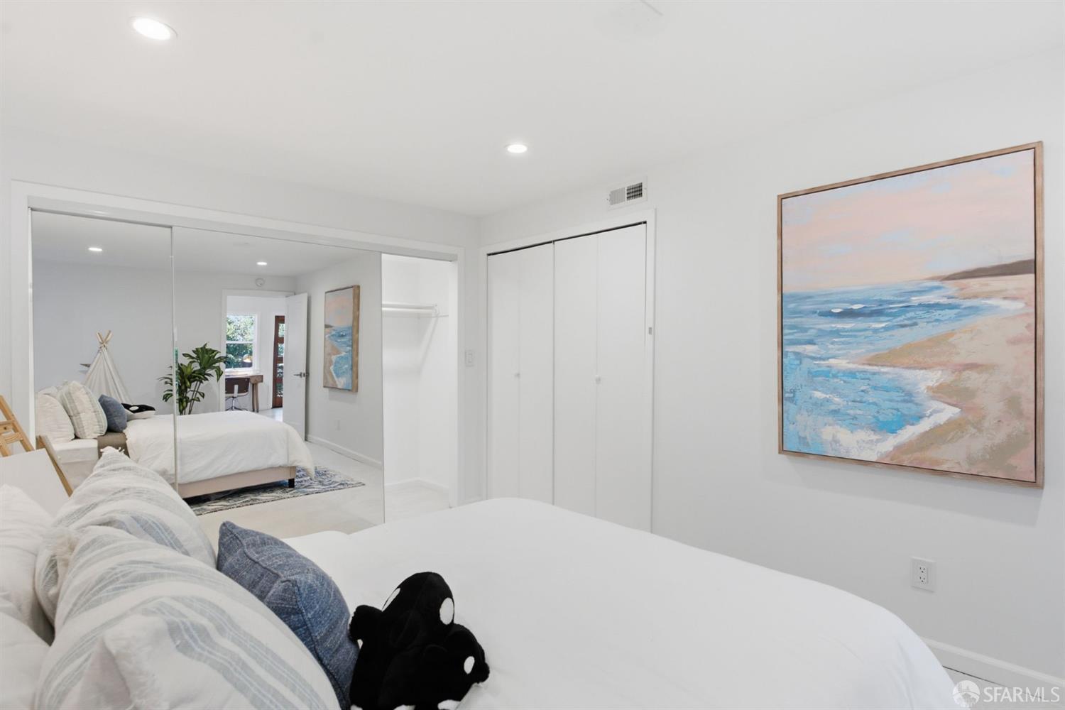 Bright bedroom with white bedding, mirrored closet doors, and framed beach artwork.