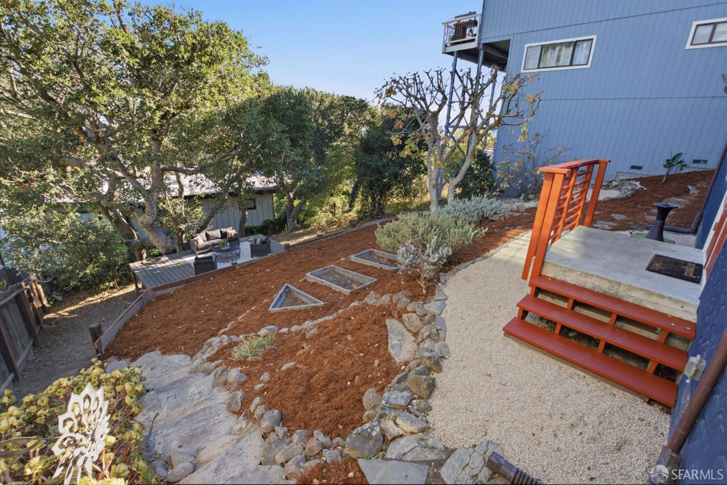 Sloped backyard with red steps, gravel paths, raised beds, and a shaded deck.