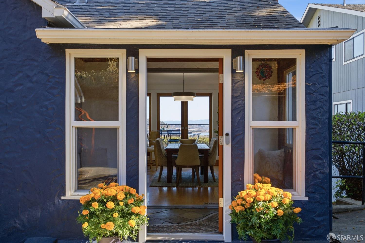 Blue home entrance with open door, orange flowers, and a glimpse of water beyond the dining room.