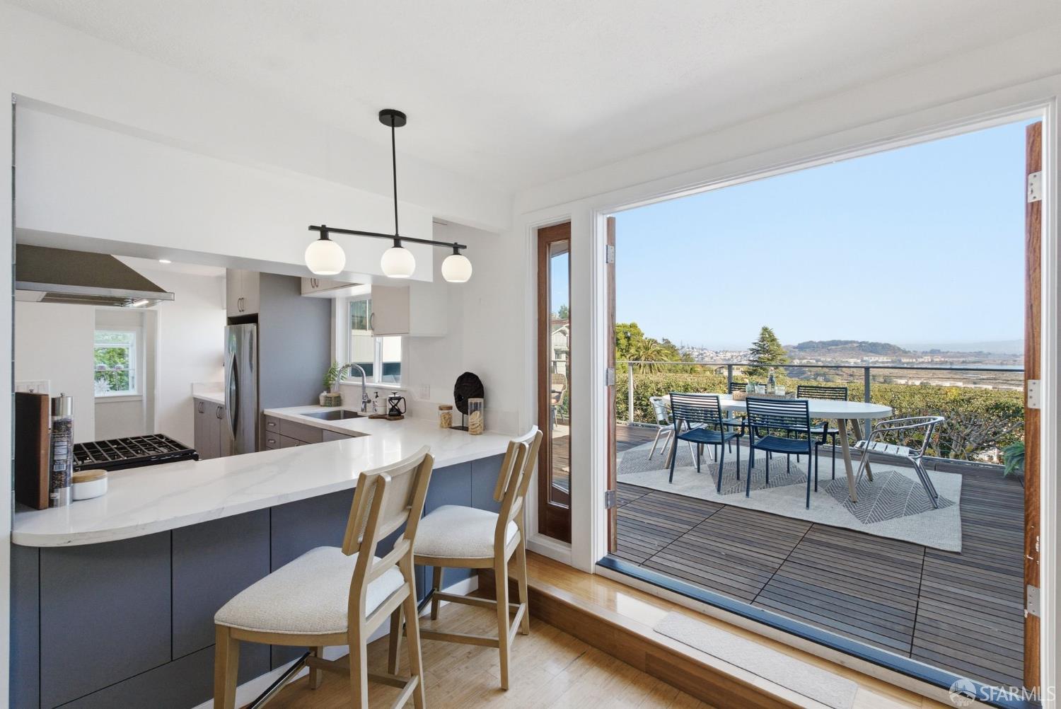Modern kitchen with a white island opens onto a deck overlooking water and hills.