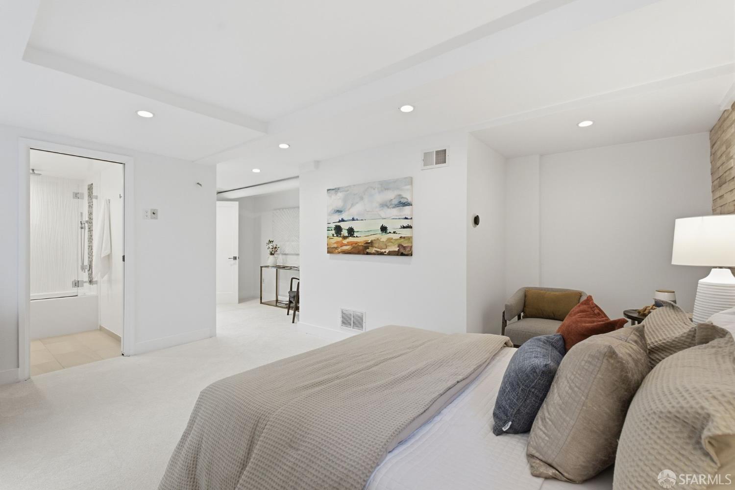 Staged bedroom with neutral bedding, a seating area, and an adjoining bathroom.