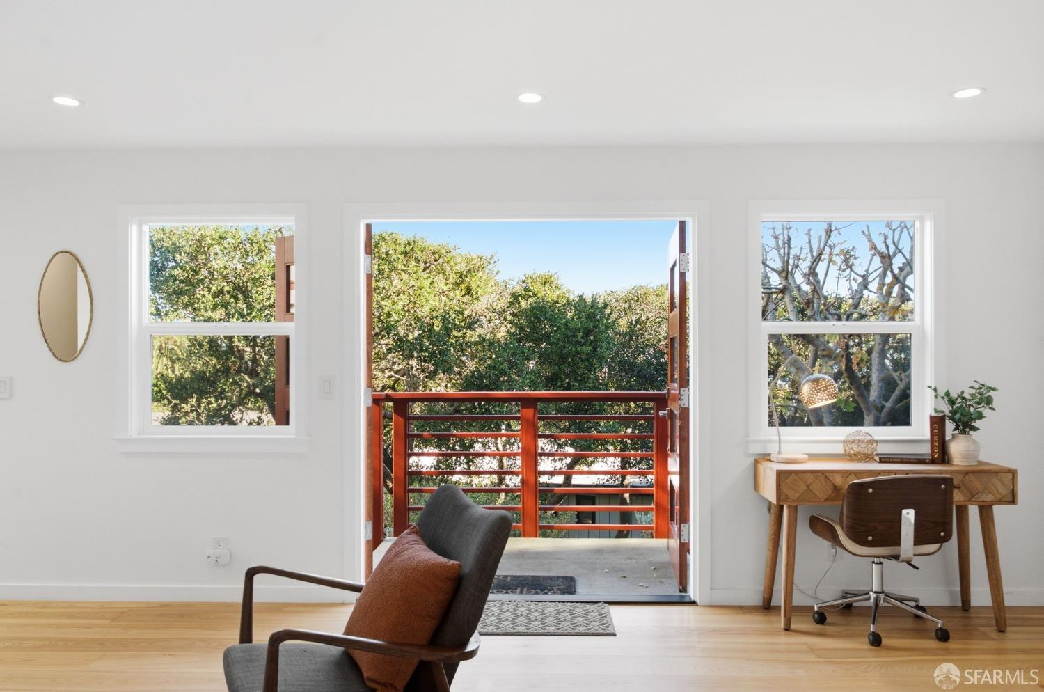 Bright room with a lounge chair, wooden desk, and open balcony doors overlooking trees.
