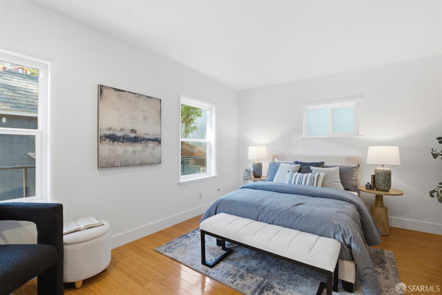 Staged bedroom with a blue bed, upholstered bench, hardwood floors, and abstract wall art.