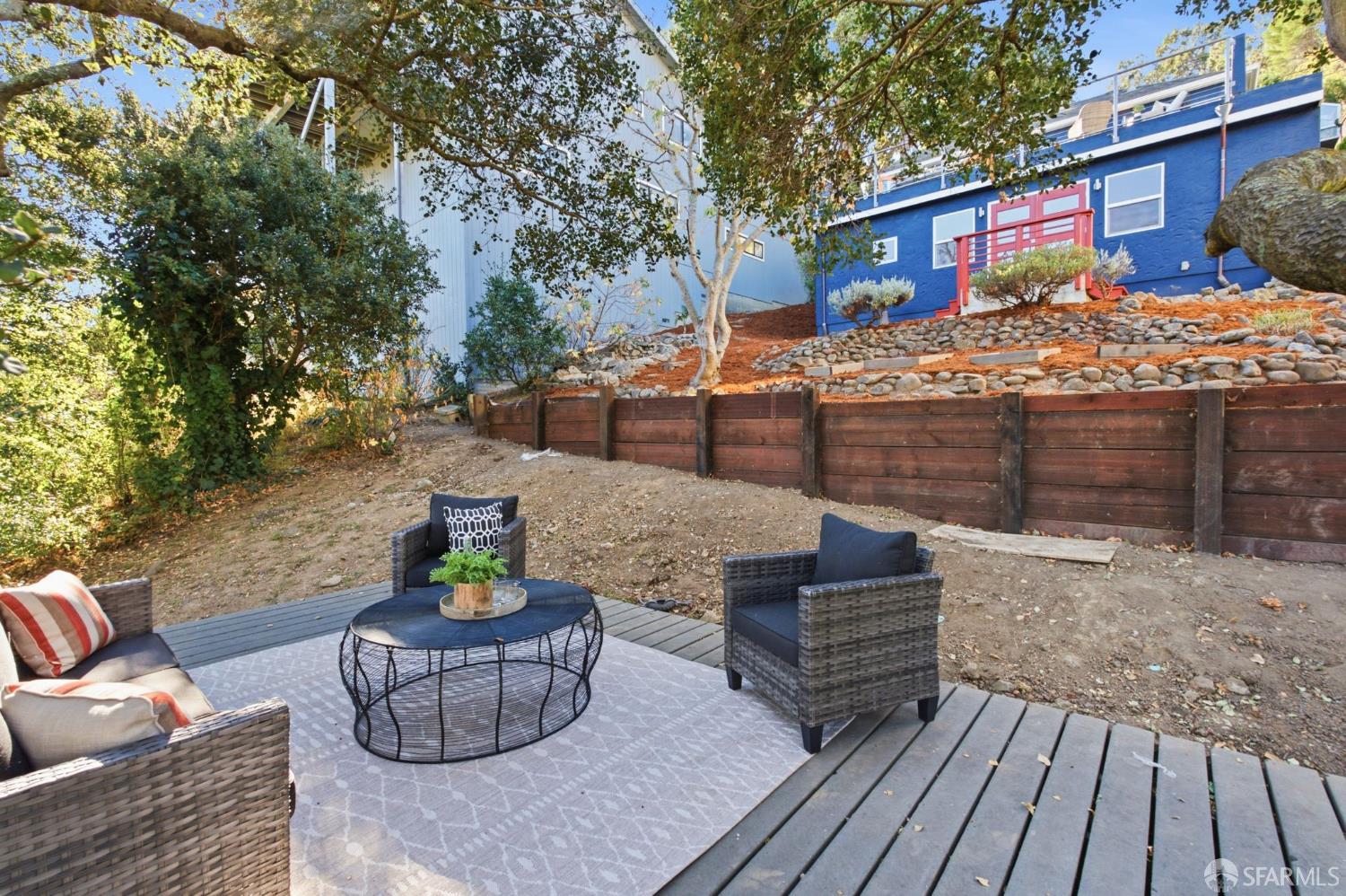 Furnished backyard deck and patio seating below a blue house on a rocky slope.