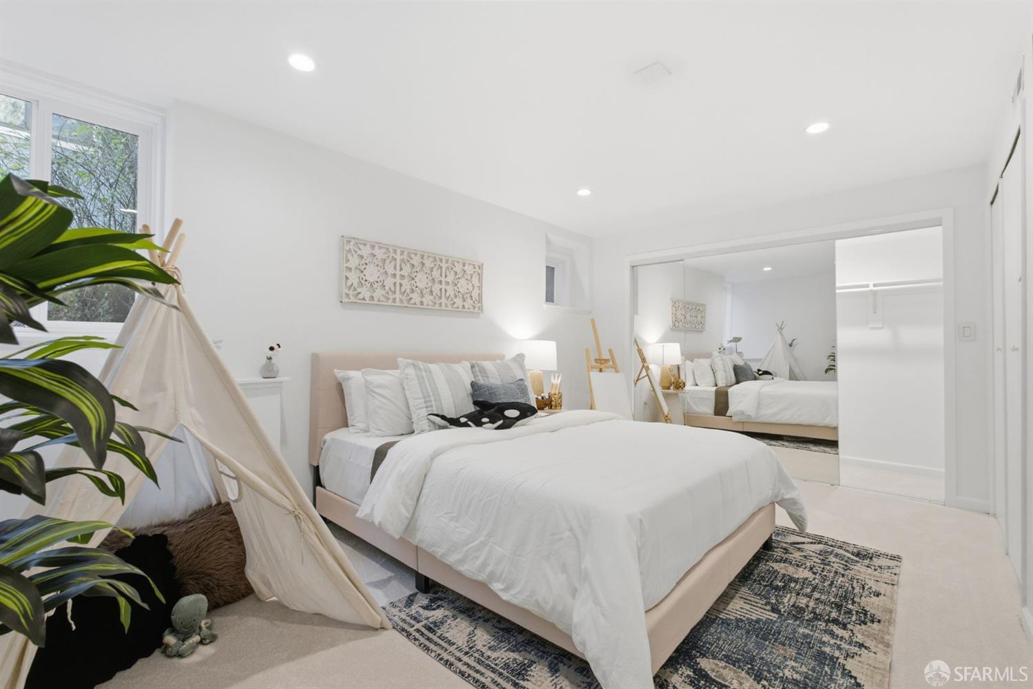 Staged bedroom with a white bed, patterned rugs, full-length mirror, and fabric teepee beside a leafy plant.