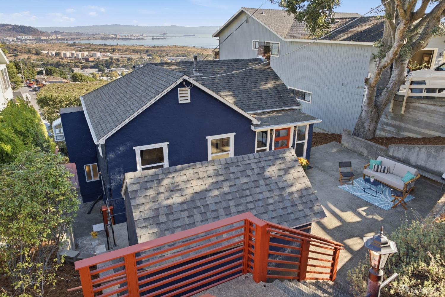 Elevated view of a dark blue house with orange-red railings, a patio, and a bay in the distance.