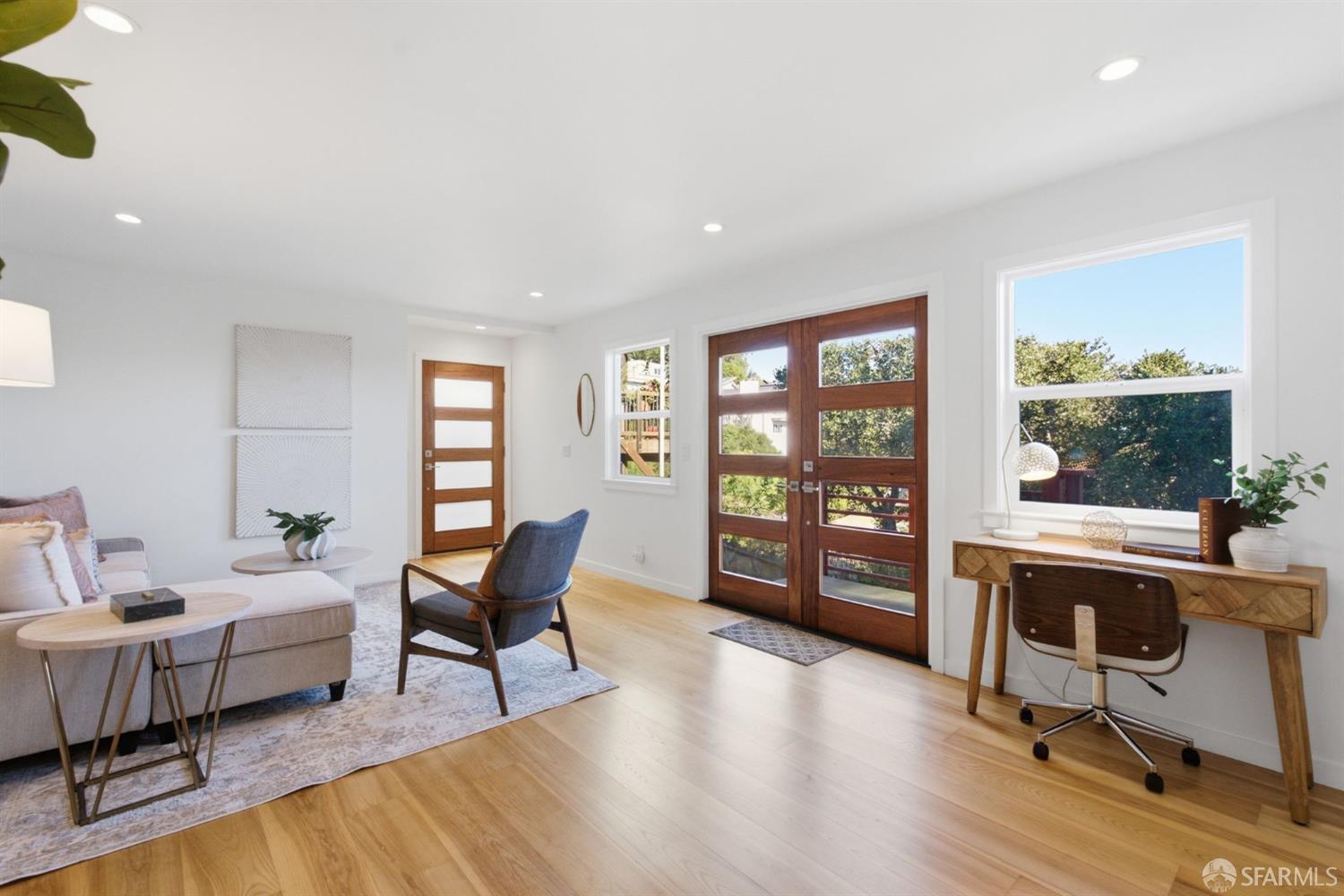 Living room with wood-framed glass doors, pale wood floors, and a desk beside a large window.