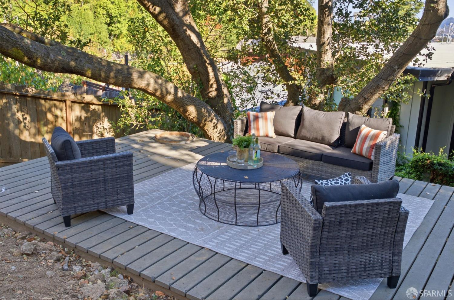 Shaded deck patio with wicker lounge seating, striped cushions, and a round coffee table beneath mature trees.