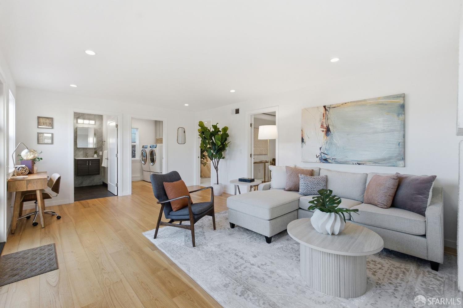Staged living room with a gray sectional, pale wood floors, and open doorways to adjacent rooms.