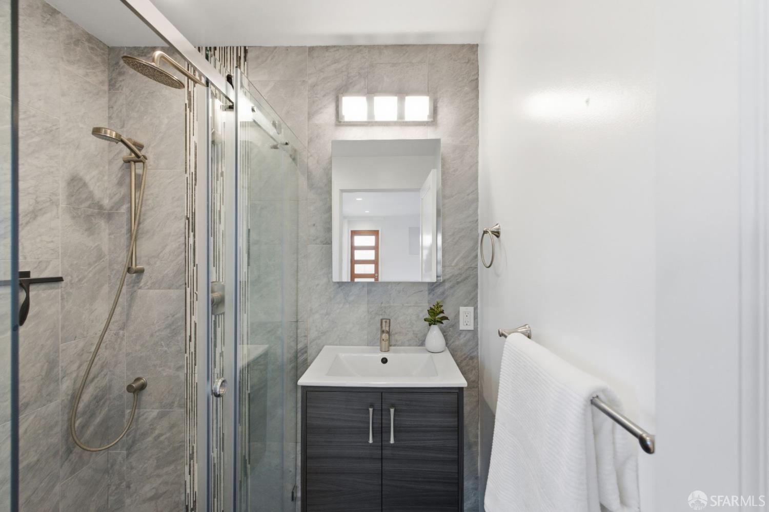 Modern bathroom with gray tiled shower, glass enclosure, and dark vanity beneath a lit mirror.