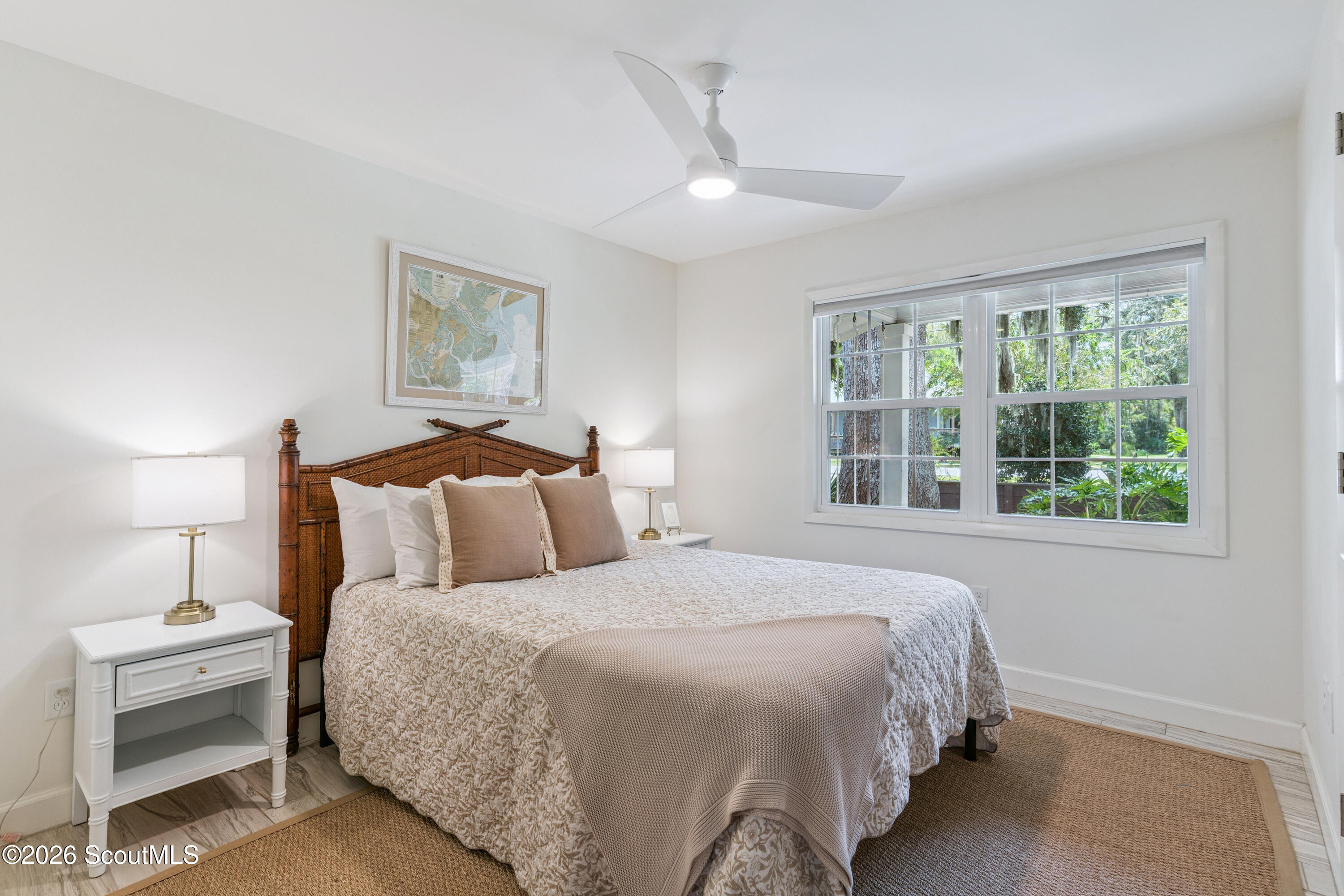 Staged bedroom with a woven wood headboard, neutral bedding, and a wide window overlooking trees.