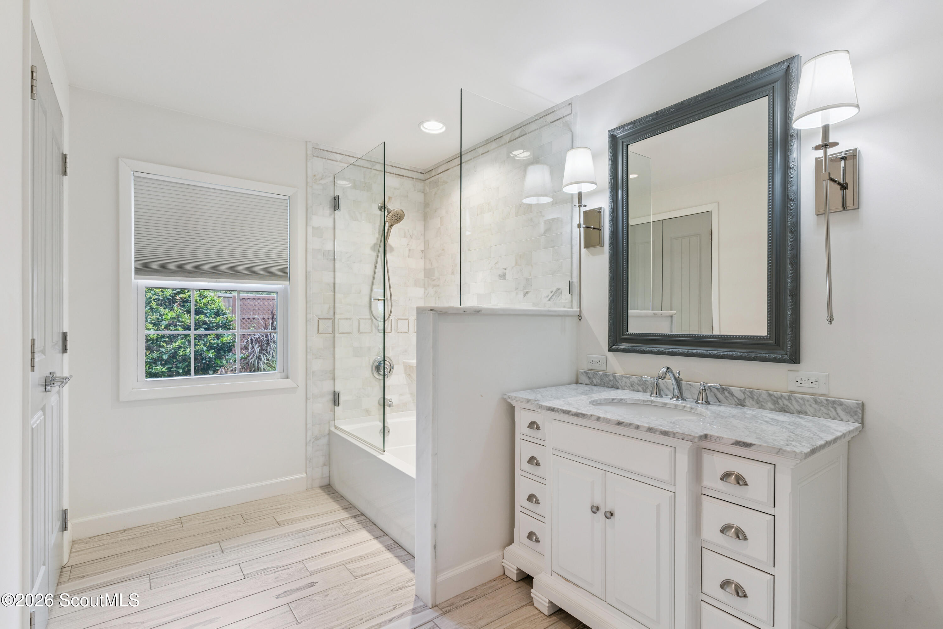 Bathroom with marble-topped vanity and glass-enclosed tub and shower beside a window overlooking greenery.