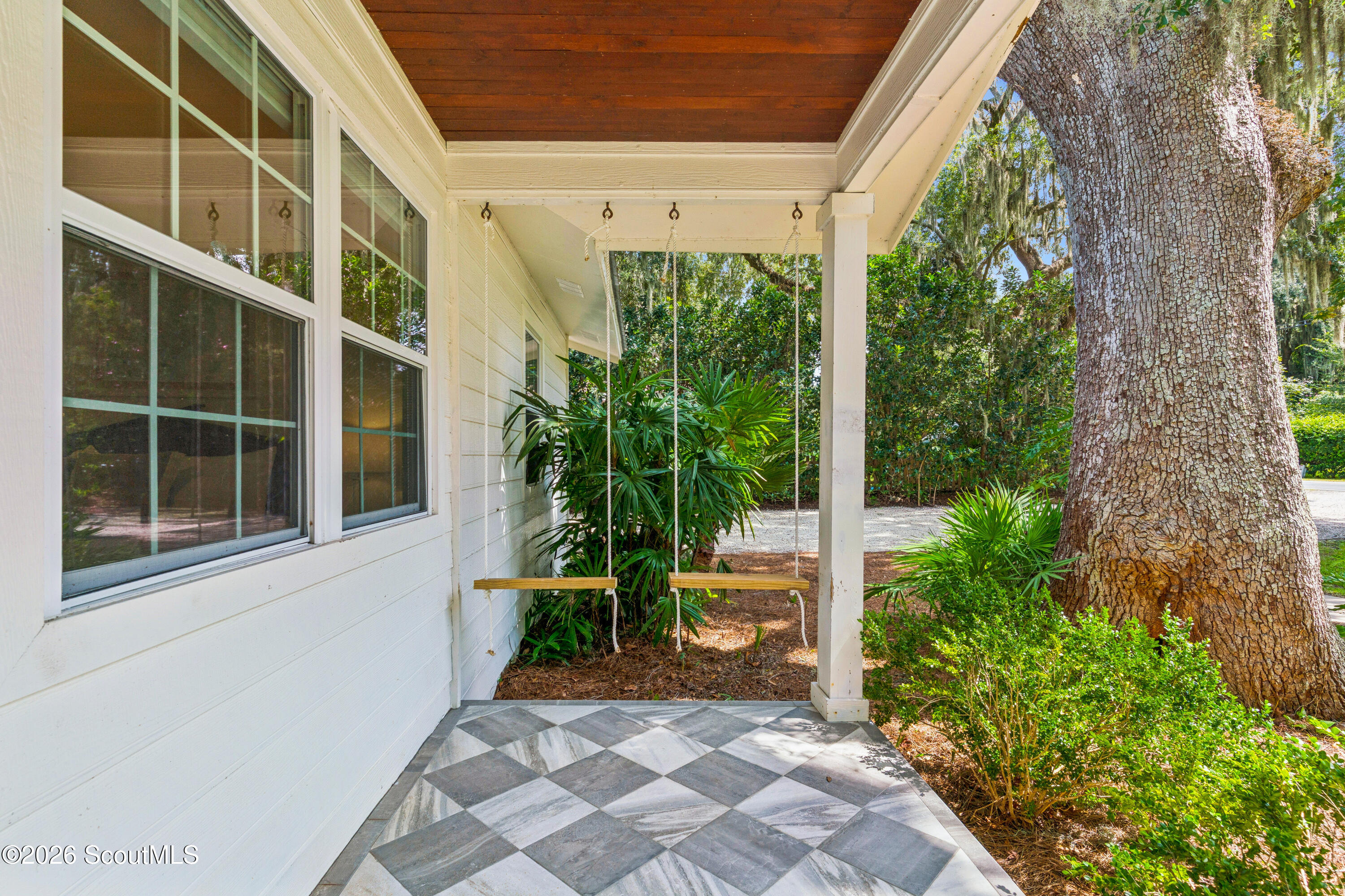 Covered porch with two wooden swings, geometric tile flooring, and a large tree beside the house.