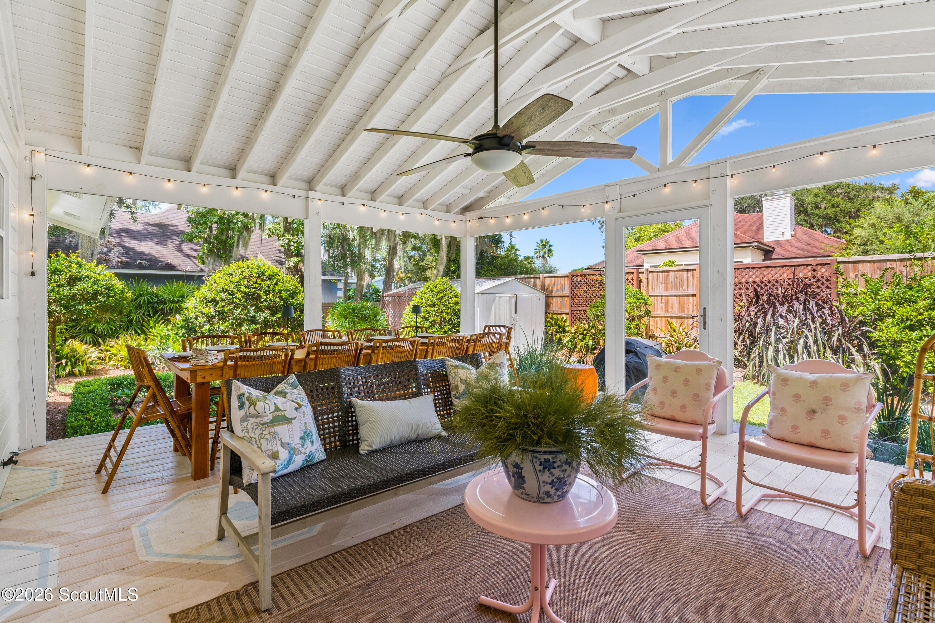 Covered patio with dining table, woven sofa, pink chairs, and a leafy fenced garden.
