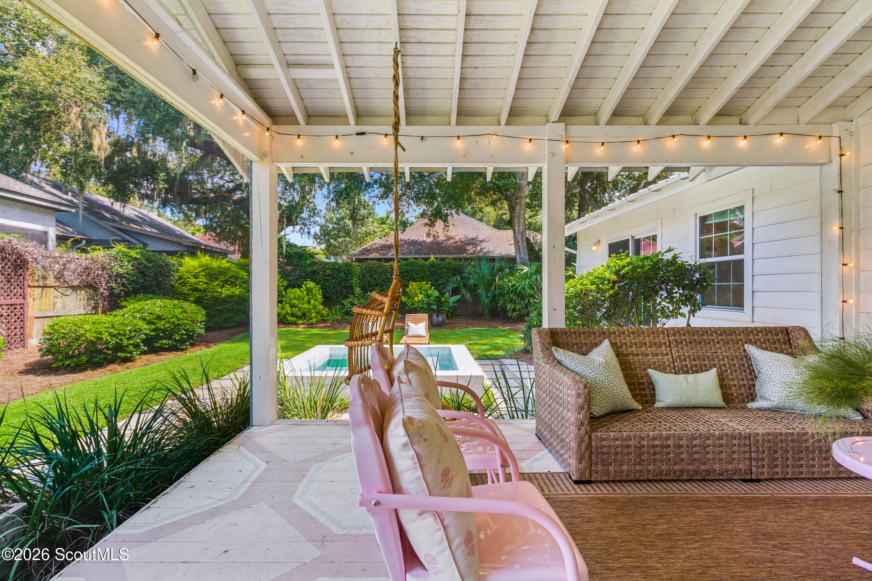 Covered backyard porch with pink chairs, wicker sofa, string lights, and a small pool beyond.