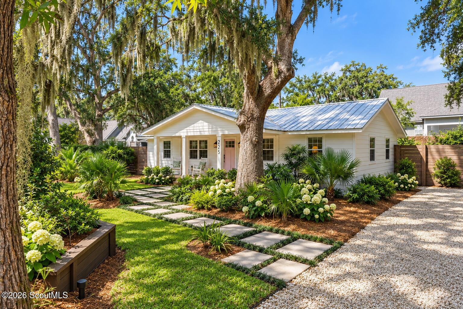 White single-story home with a pink front door, metal roof, lush landscaping, and a stepping-stone walkway beneath a moss-draped oak.