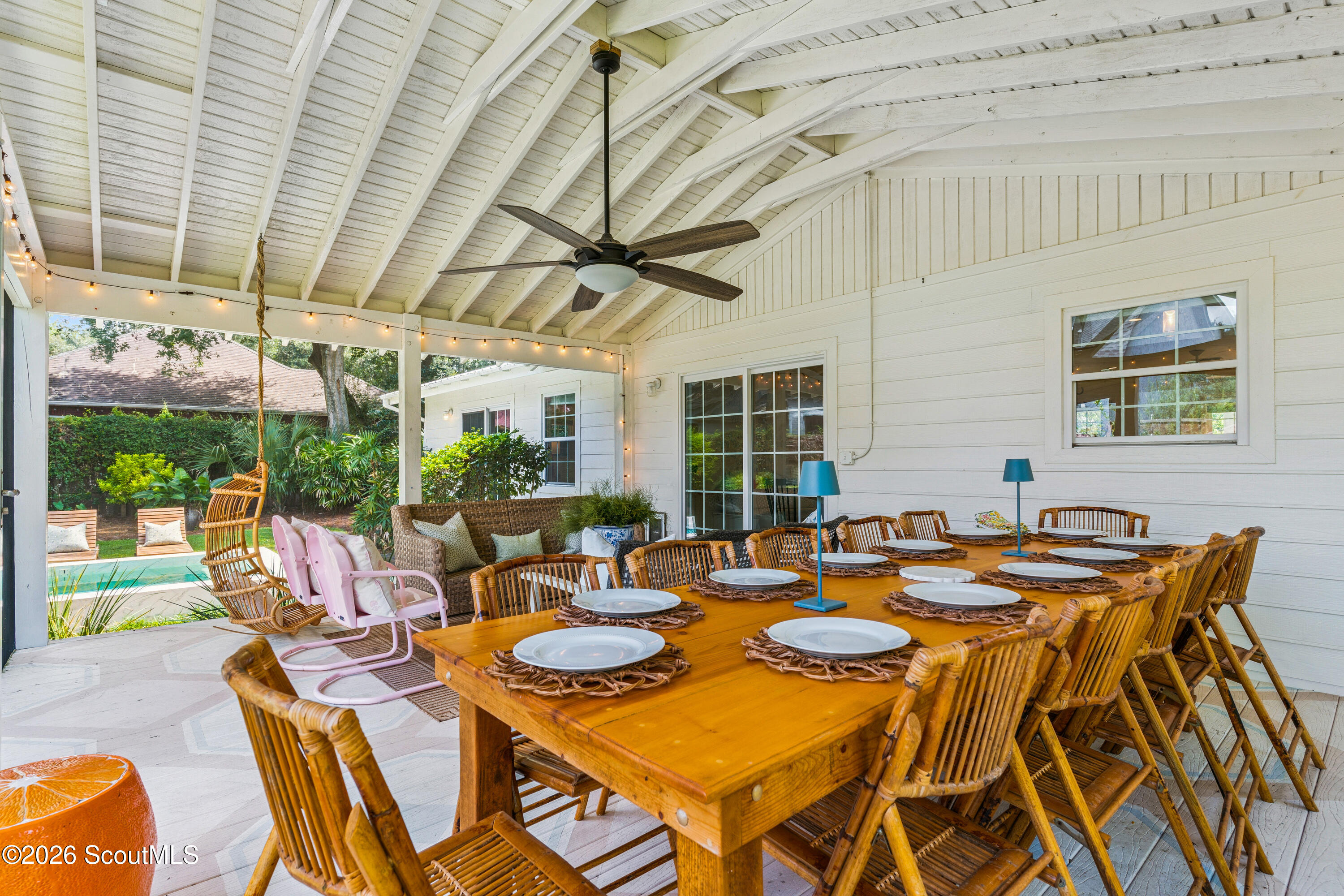 Covered dining porch with a long wooden table, bamboo chairs, and a view of the pool and garden.