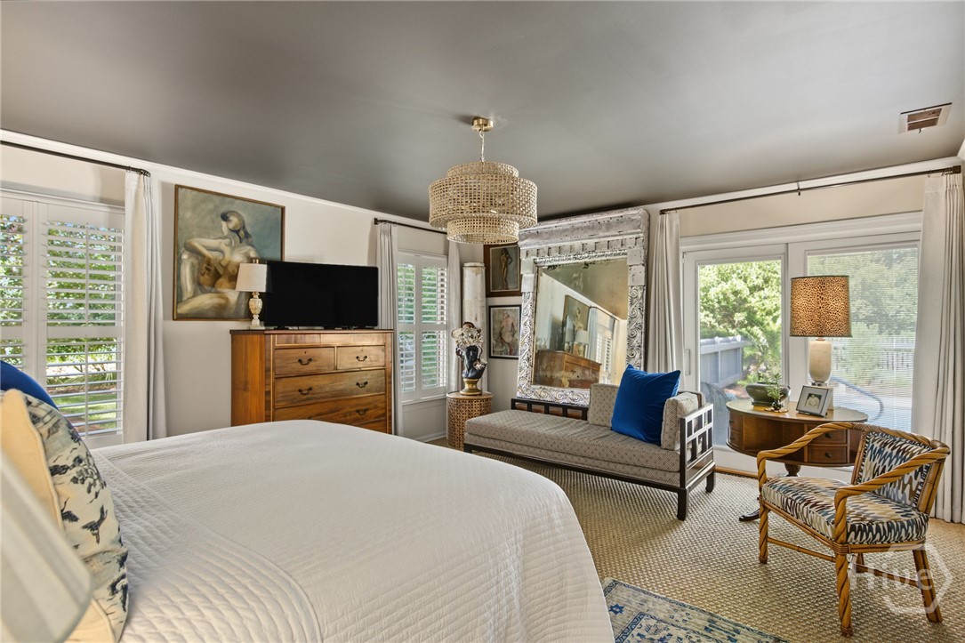 Furnished bedroom with a large white bed, ornate mirror, and sitting area beside garden-facing glass doors.