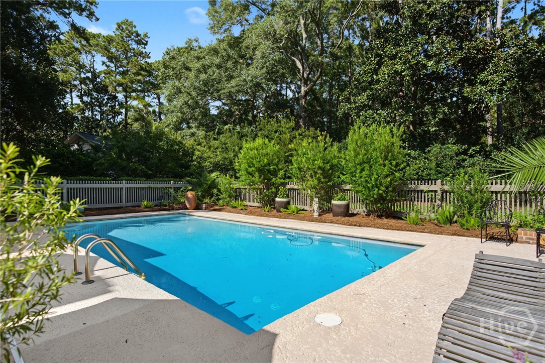 Swimming pool with a pale stone patio, white fence, and leafy trees.