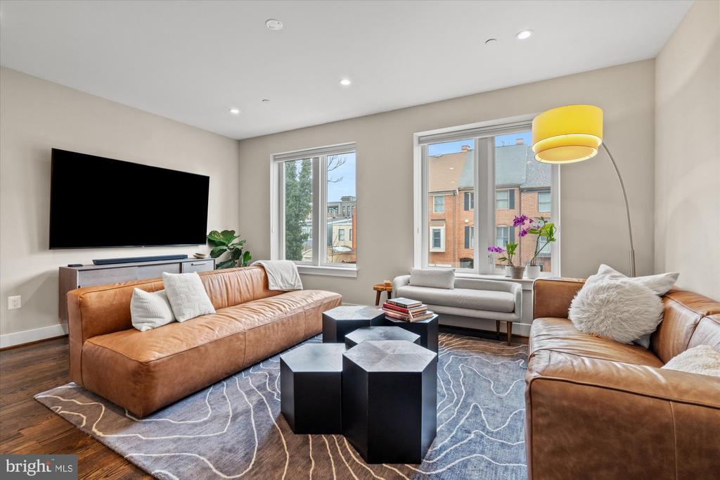 Living room with tan leather sofas, geometric coffee tables, and windows overlooking neighboring townhouses.