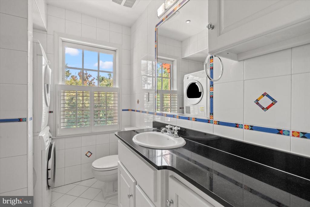 Bathroom with black countertop, colorful tile accents, shuttered window, and stacked washer and dryer.