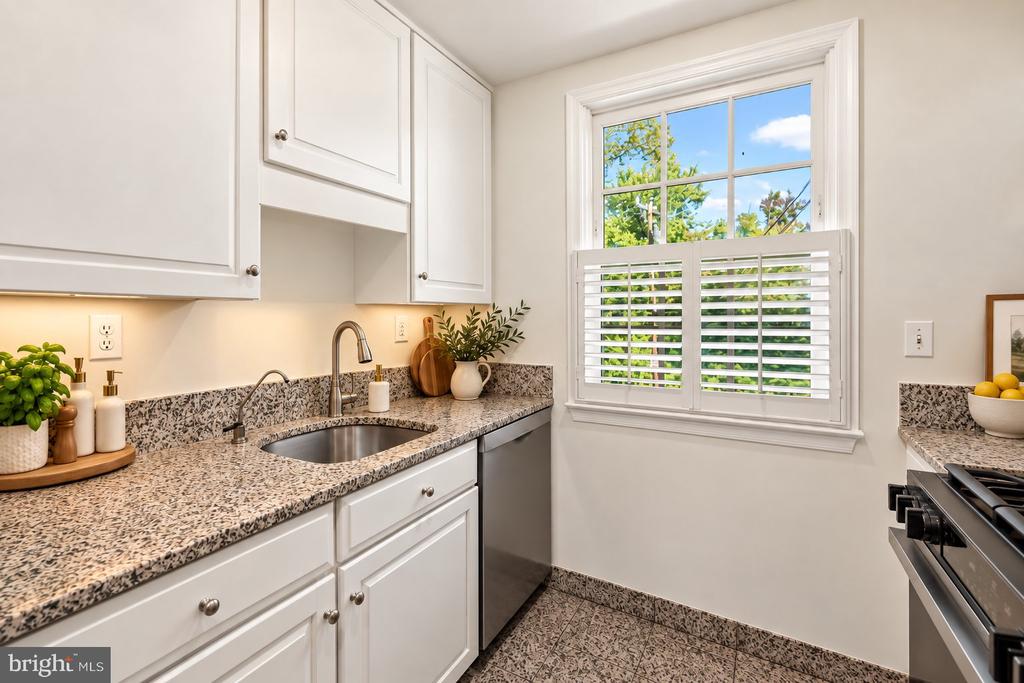 Bright kitchen with white cabinets, speckled countertops, and a shuttered window overlooking greenery.