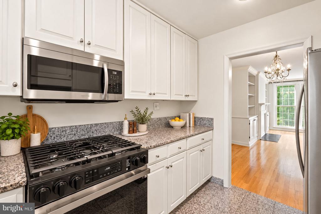White kitchen with a black gas range, stainless microwave, speckled counters, and doorway to a bright hardwood-floored room.