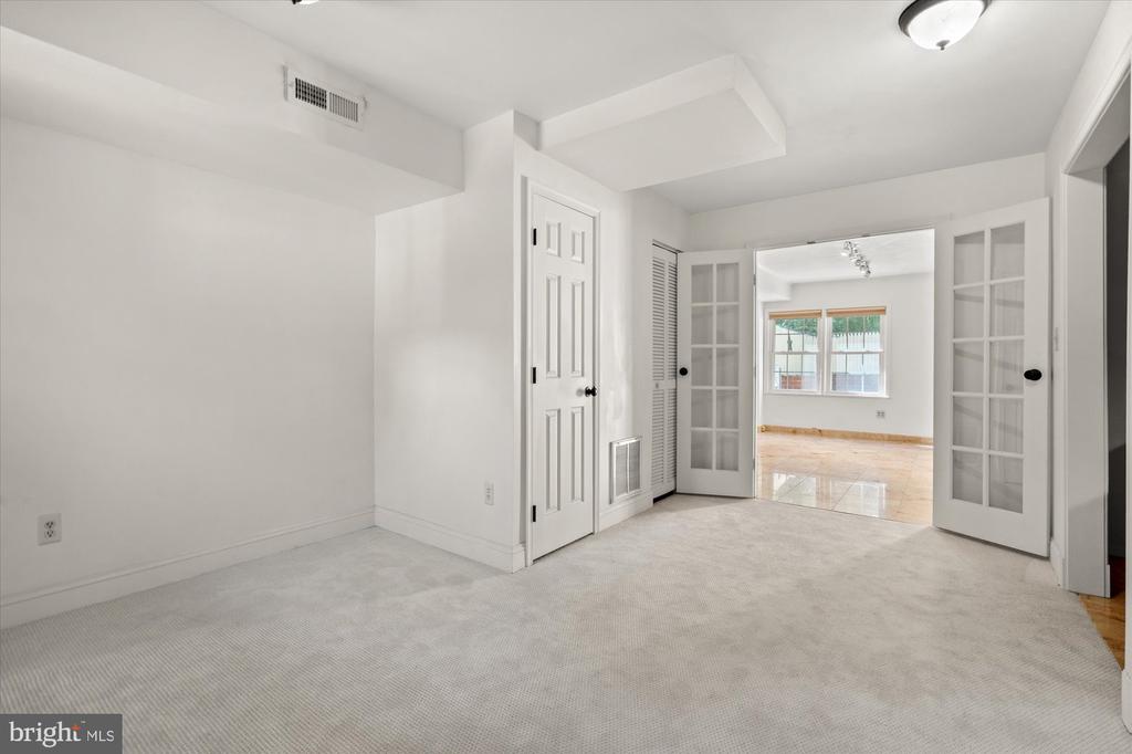 Empty carpeted room with open French doors leading to a bright room with hardwood floors.