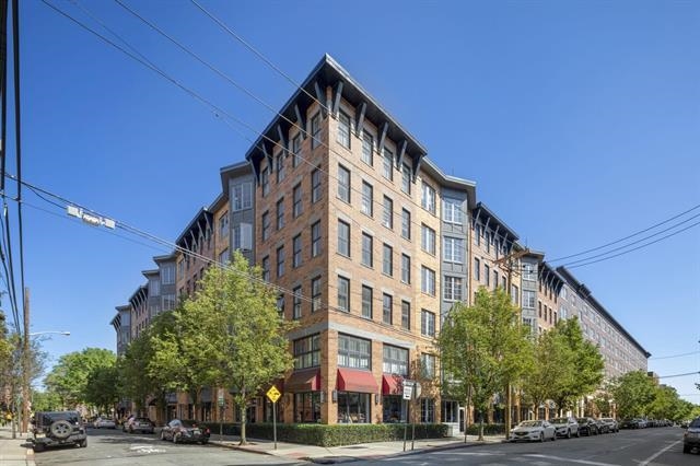 Multi-story brick corner building with storefronts and leafy trees along an urban street.