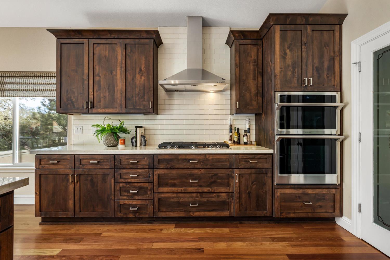 Kitchen with dark wood cabinetry, white tile backsplash, gas cooktop, and double wall ovens.