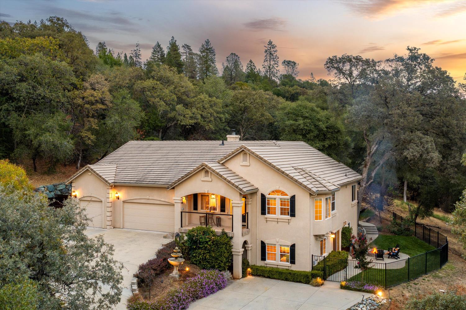 Two-story beige stucco home with a tile roof, three-car garage, and wooded hillside behind it.