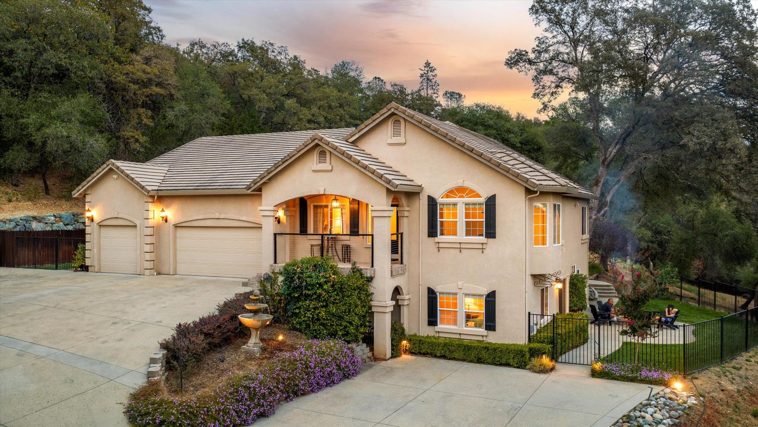 Two-story beige house with a three-car garage, lit windows, and wooded surroundings at dusk.