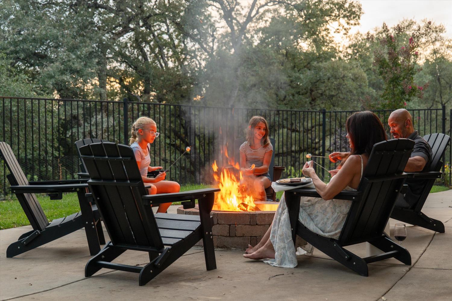 Backyard patio with Adirondack chairs around a stone fire pit as people roast marshmallows.