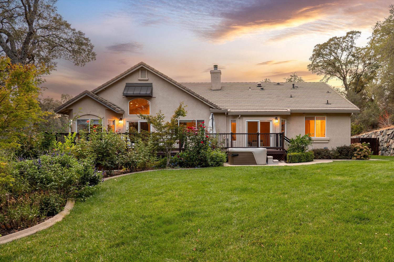 Home’s rear exterior with a deck, hot tub, landscaped garden, and broad lawn at sunset.