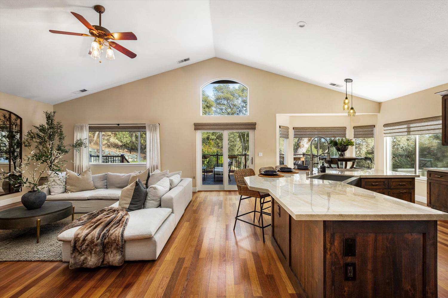 Open-plan kitchen and living room with a vaulted ceiling, stone island, hardwood floors, and large windows.