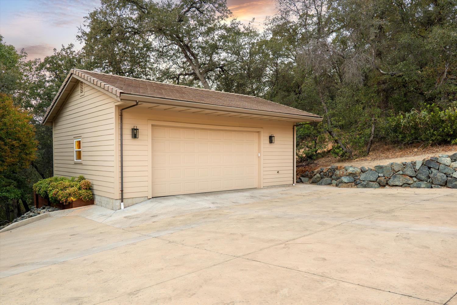 Beige detached two-car garage with a broad concrete driveway beneath mature trees.
