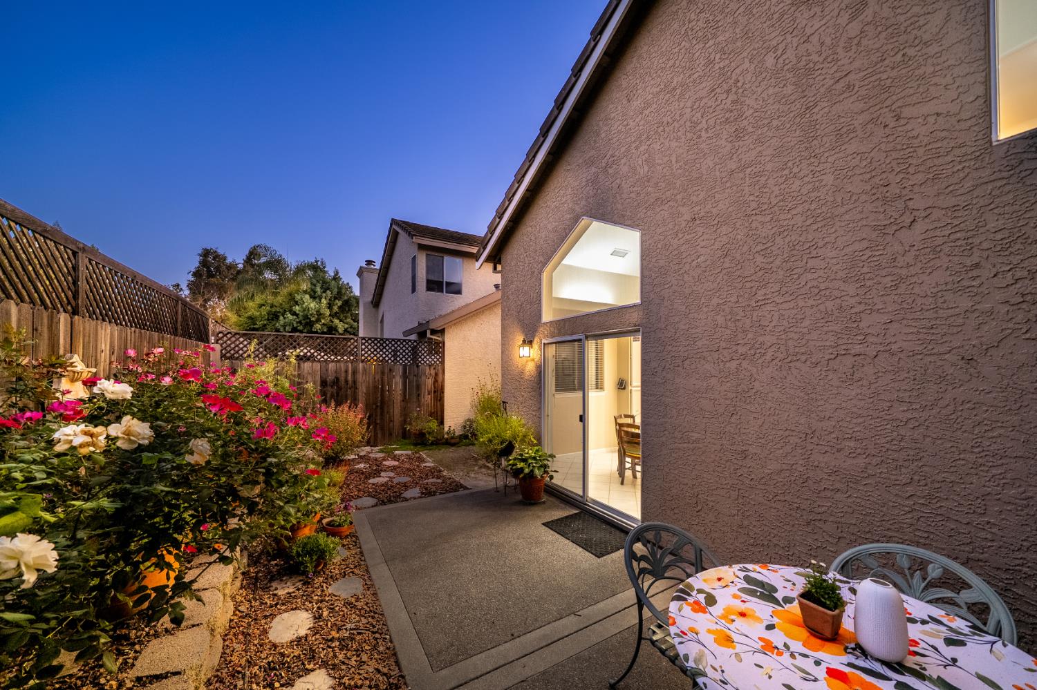 Back patio with outdoor dining set beside a stucco home and colorful flower garden.