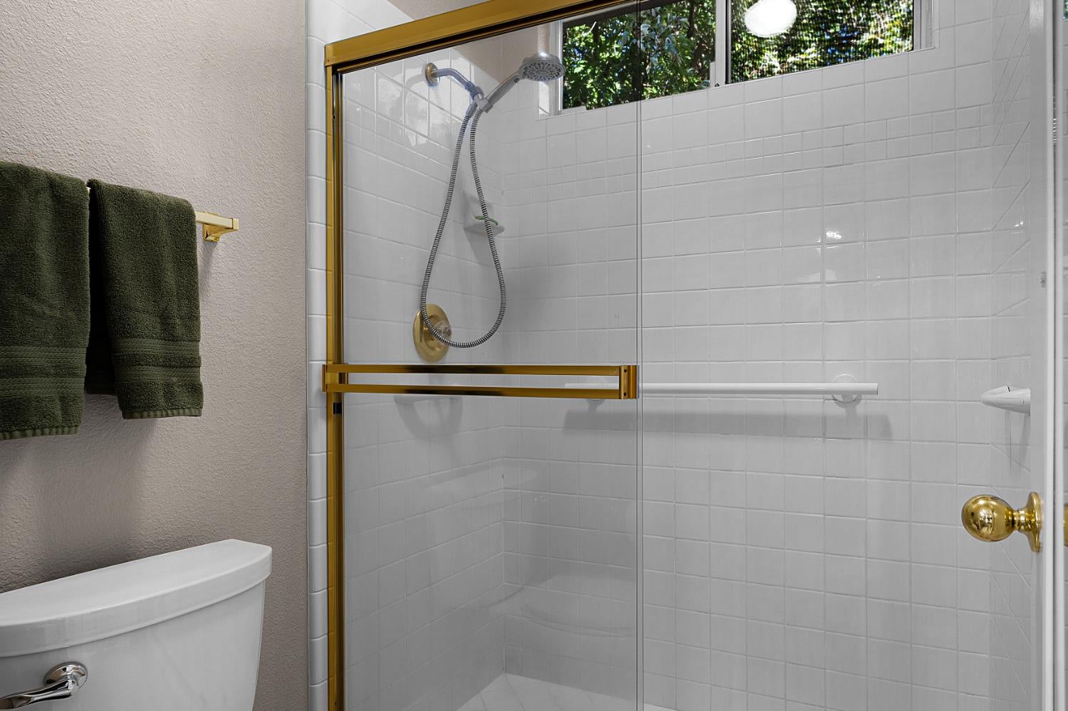 White-tiled glass shower with gold-tone trim and handheld showerhead beside dark green towels.