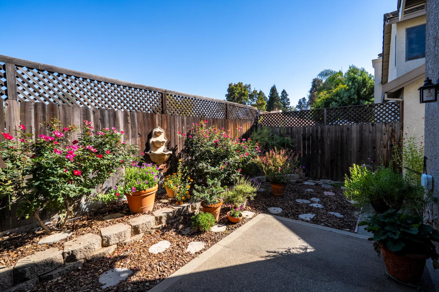 Landscaped backyard patio with flowering potted plants, stepping stones, and a wall fountain.