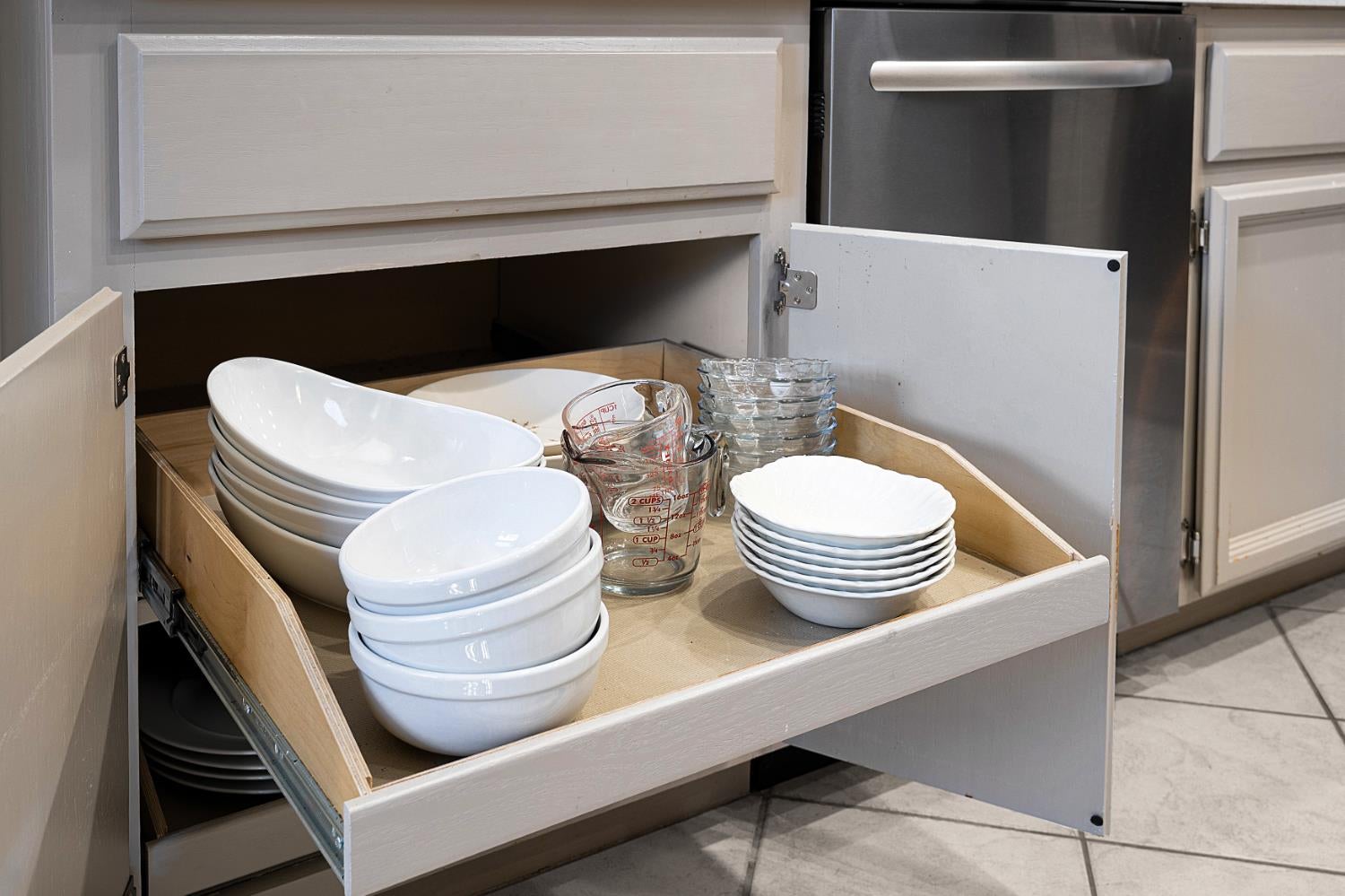Open kitchen drawer stocked with dishes and measuring cups beside a stainless-steel dishwasher.