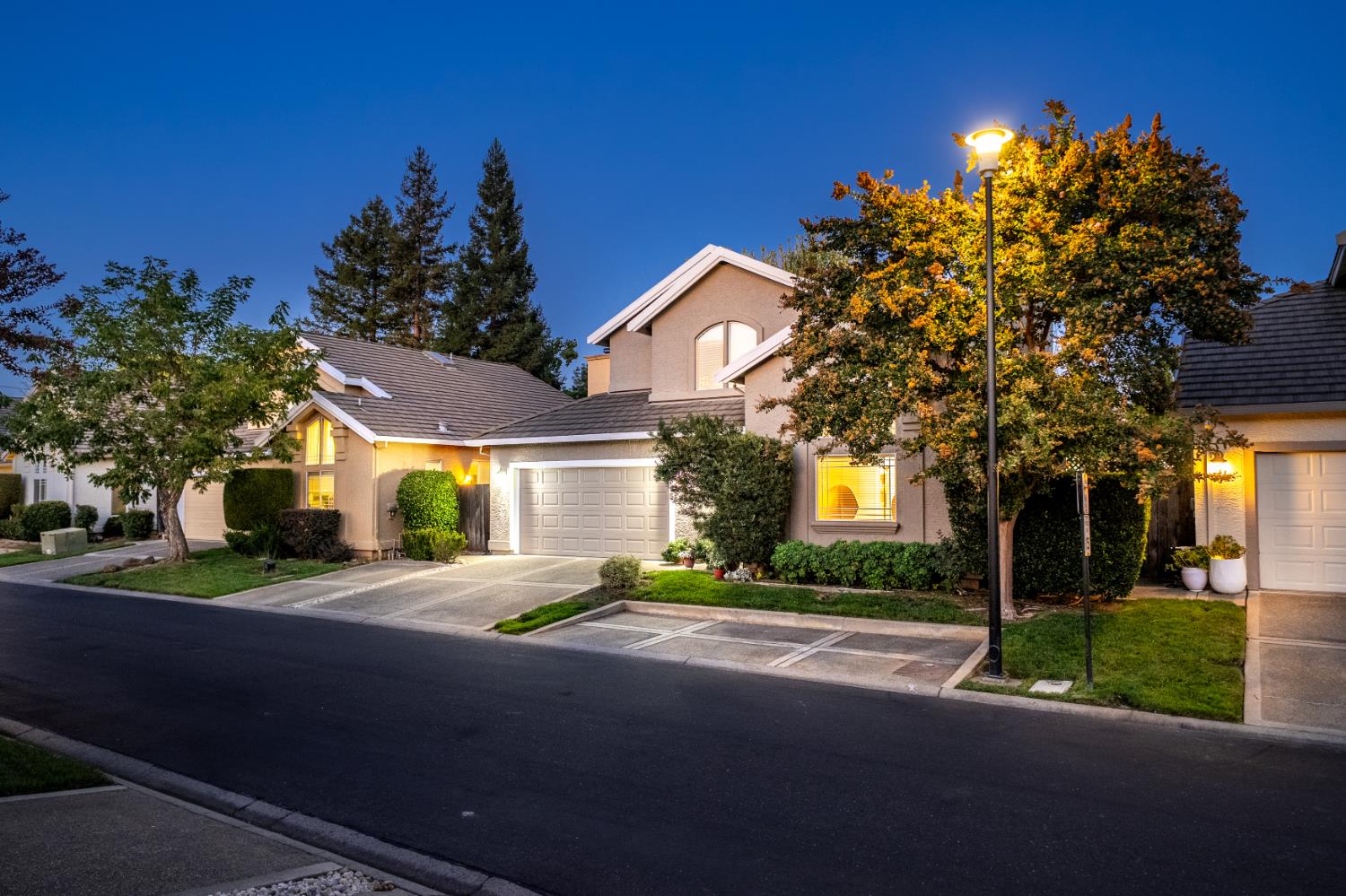 Two-story beige home with a two-car garage and autumn trees, photographed at dusk.